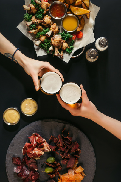 partial top view of people drinking beer and eating assorted meat and baked potatoes with roasted chicken on black - Фото, изображение