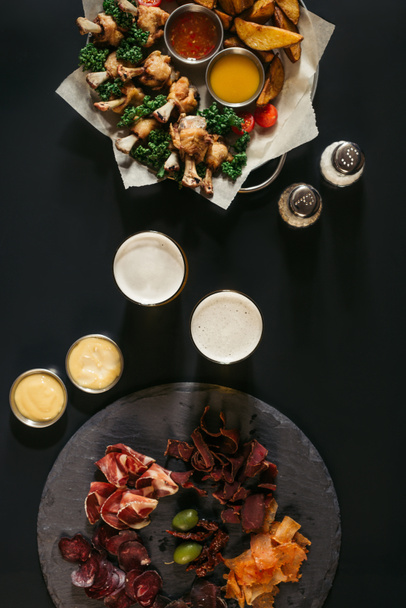 pohled shora sklenic piva, různé maso a smažené brambory s kuřetem na černém pozadí - Fotografie, Obrázek