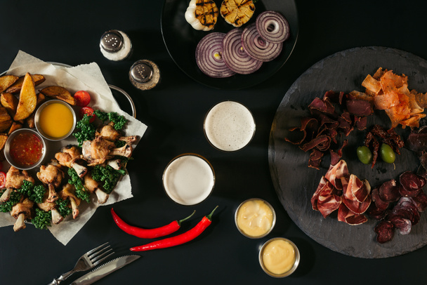 vista superior de copos de cerveja, legumes, batatas assadas com frango assado, vários molhos e carne artesanal em preto
 - Foto, Imagem