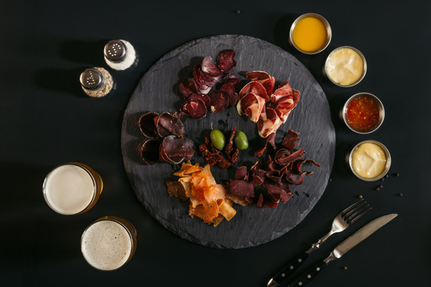 top view of gourmet assorted meat on slate board, various sauces and glasses of beer on black  - Photo, Image