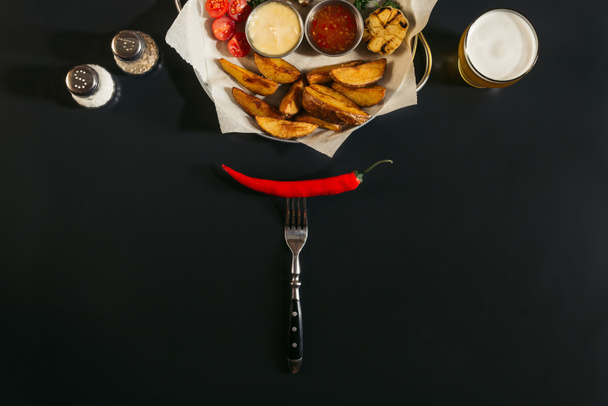 top view of red hot chili pepper on fork and delicious roasted potatoes with sauces on black - Photo, Image