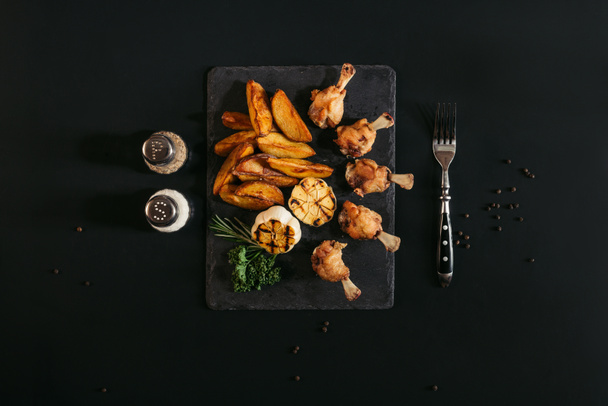bovenaanzicht van heerlijke geroosterde aardappelen met kip en gegrilde knoflook op leisteen bord op zwart  - Foto, afbeelding