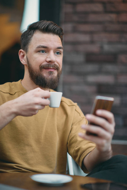 Молодой человек сидит в кафе и пользуется смартфоном
. - Фото, изображение