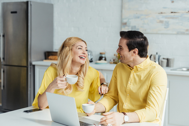 Happy couple drinking coffee and talking by table with laptop - Фото, изображение