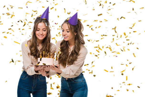 smiling twins in birthday caps looking at birthday cake under falling confetti isolated on white - Foto, immagini