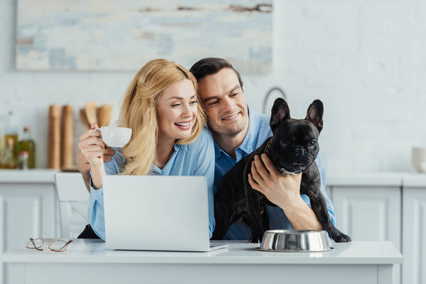 Man and woman drinking coffee and hugging their dog on kitchen table with laptop - Zdjęcie, obraz