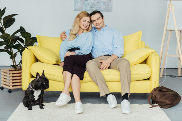 Bonito casal sentado no sofá amarelo por bulldog francês no chão
 - Foto, Imagem