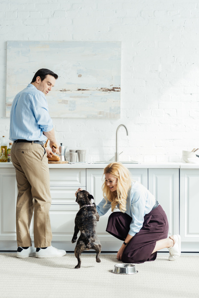 Man preparing food and woman playing with french bulldog on kitchen floor - Фото, изображение