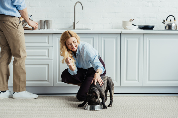 Homem cervejando café e mulher loira acariciando bulldog francês no chão da cozinha
 - Foto, Imagem