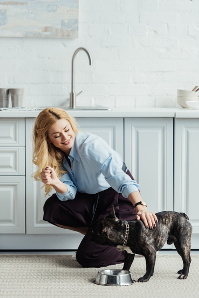 Uśmiechnięta Blondynka głaskanie frenchie psa na podłodze w kuchni - Zdjęcie, obraz