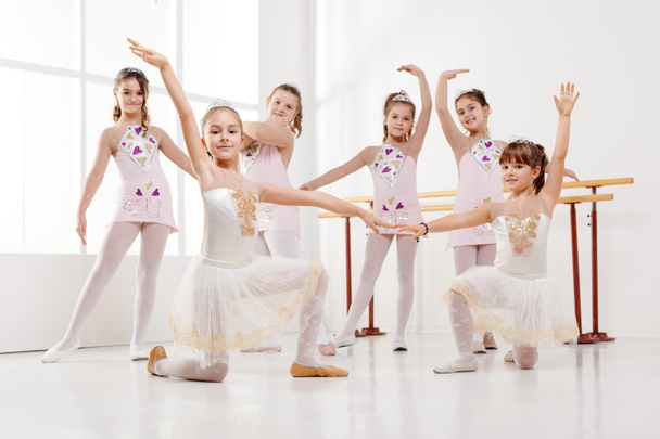 Улыбающиеся маленькие девочки в платьях, практикующие позы во время балетных занятий. Смотреть в камеру
. - Фото, изображение