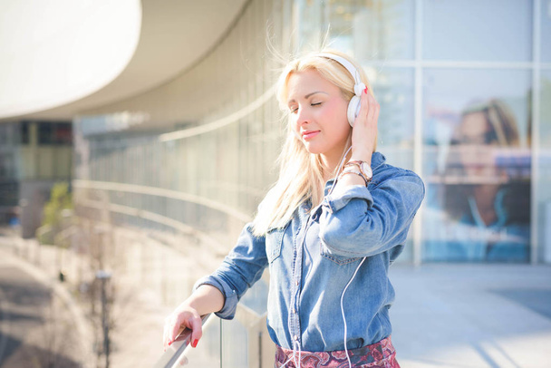 Fél hosszúságú, egy fiatal gyönyörű kaukázusi szőke lány hajol egy ablakpárkányon fejhallgató a város zenét hallgat - Fotó, kép