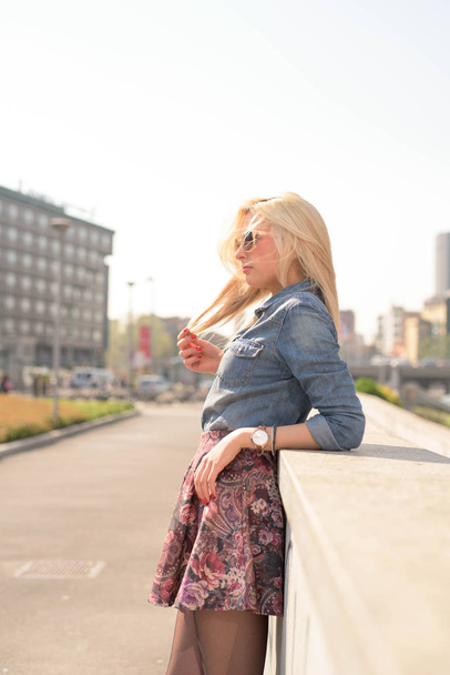 Nuori kaunis blondi valkoihoinen tyttö poseeraa ulkona kaupungissa yllään farkut paita ja kukka hame katselee pois yllään aurinkolasit
 - Valokuva, kuva