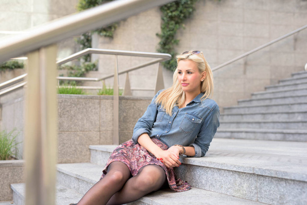 Young beautiful blonde caucasian girl sitting posing outdoor in the city wearing a jeans shirt and a floral skirt looking away - Fotoğraf, Görsel