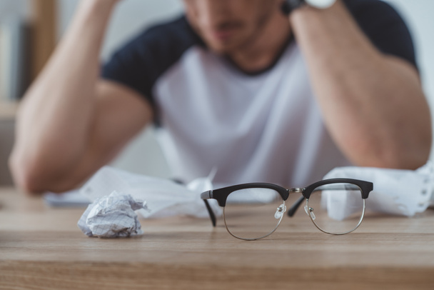 selective focus of depressed student at table with crumpled papers and eyeglasses on foreground - Photo, Image