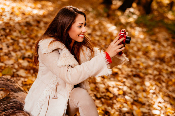 Beautiful smiling girl enjoying in forest in autumn. She is sitting on the tree and taking photo for memory. - 写真・画像