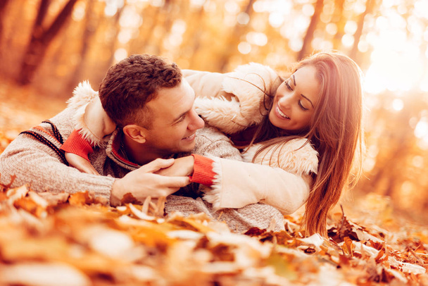 Belo casal sorridente desfrutando na floresta ensolarada nas cores do outono. Eles estão deitados nas folhas de quedas e se divertindo
. - Foto, Imagem
