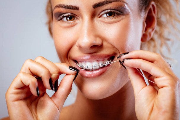 Крупный план улыбающейся девушки с брекетами для чистки зубов зубной нитью. Смотреть в камеру
. - Фото, изображение