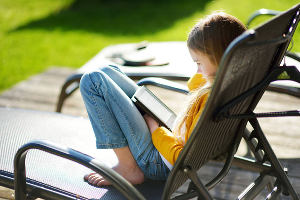 Cute little girl sitting in a garden and reading an ebook on sunny summer evening. Education for kids. - Photo, Image