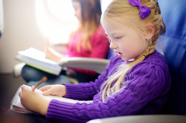 Αξιολάτρευτο μικρό κορίτσια ταξιδεύουν με αεροπλάνο. Το παιδί κάθεται από αεροσκάφη παράθυρο και σχεδιάζοντας μία εικόνα με πολύχρωμα μολύβια. Ταξίδια στο εξωτερικό με τα παιδιά. - Φωτογραφία, εικόνα