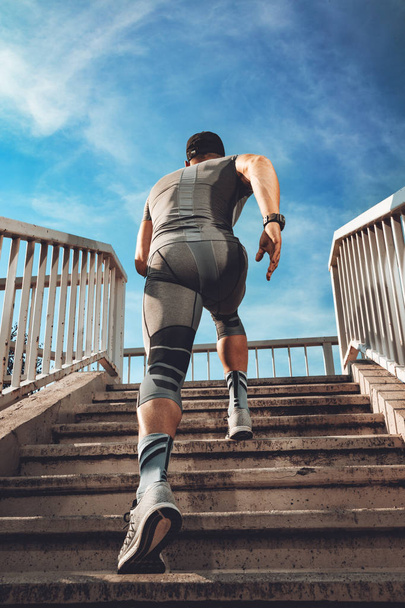 Молодой мускулистый спортсмен бежит вверх по лестнице на мосту. Вид сзади
.  - Фото, изображение