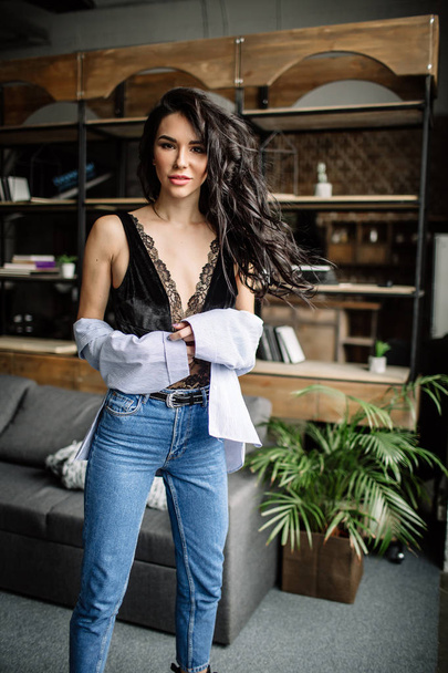Foreground. Beautiful brunette dressed in a blue shirt and jeans in an room in the back background of a bookshelf and sofa, fashion style - Foto, Imagem