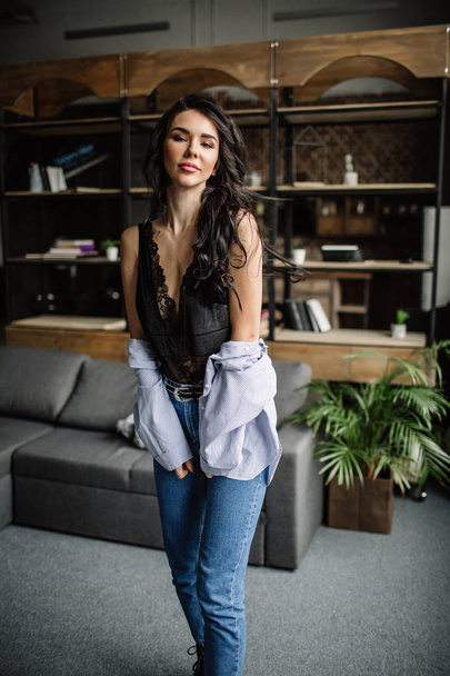Foreground. Beautiful brunette dressed in a blue shirt and jeans in an room in the back background of a bookshelf and sofa, fashion style - Foto, Bild