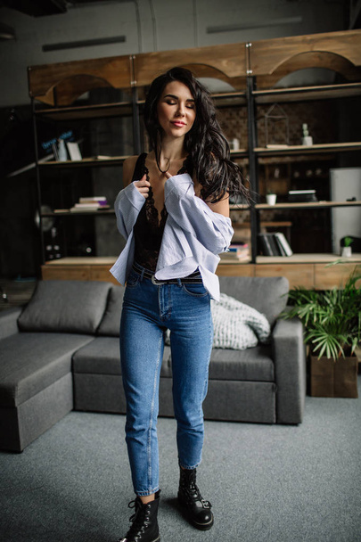 Foreground. Beautiful brunette dressed in a blue shirt and jeans in an room in the back background of a bookshelf and sofa, fashion style - Photo, Image