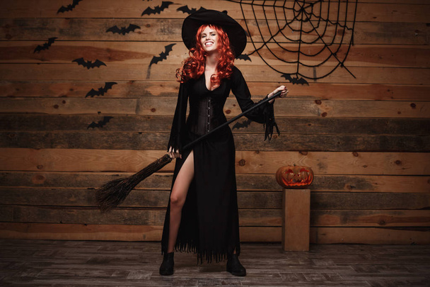 Conceito de bruxa de Halloween - Full-length Happy Halloween cabelo vermelho Witch segurando posando com vassoura mágica sobre o velho estúdio de madeira fundo
. - Foto, Imagem