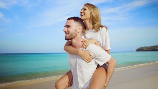 Belo casal feliz se divertindo, fazendo piggyback na praia de areia tropical sobre o fundo do mar e do céu
 - Foto, Imagem