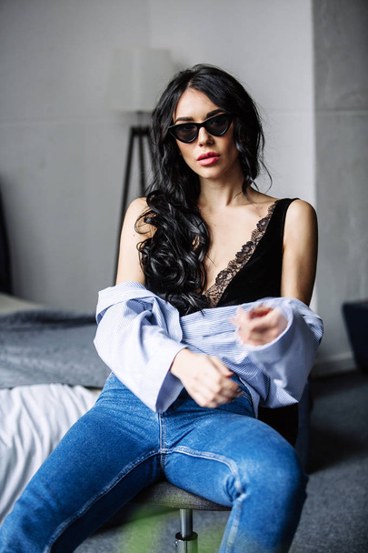 portrait of a beautiful brunette in sunglasses, blue shirt, blue jeans and interior in room - Fotó, kép