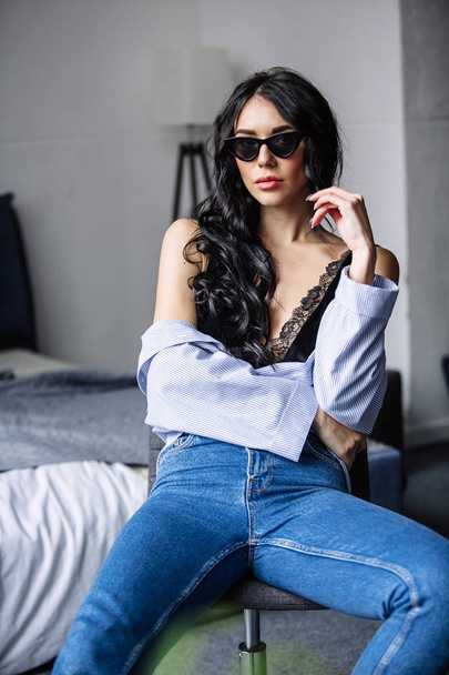 portrait of a beautiful brunette in sunglasses, blue shirt, blue jeans and interior in room - Photo, Image