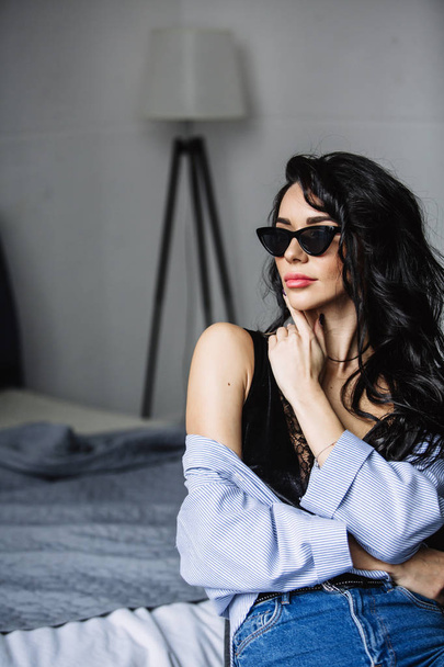 portrait of a beautiful brunette in sunglasses, blue shirt, blue jeans and interior in room - Photo, image