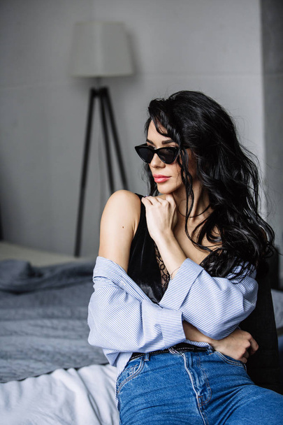 portrait of a beautiful brunette in sunglasses, blue shirt, blue jeans and interior in room - Fotó, kép