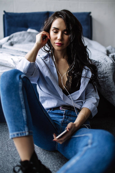 A beautiful brunette listens to music in headphones in a room near the sofa, is dressed in a shirt and blue jeans, blue - Фото, изображение