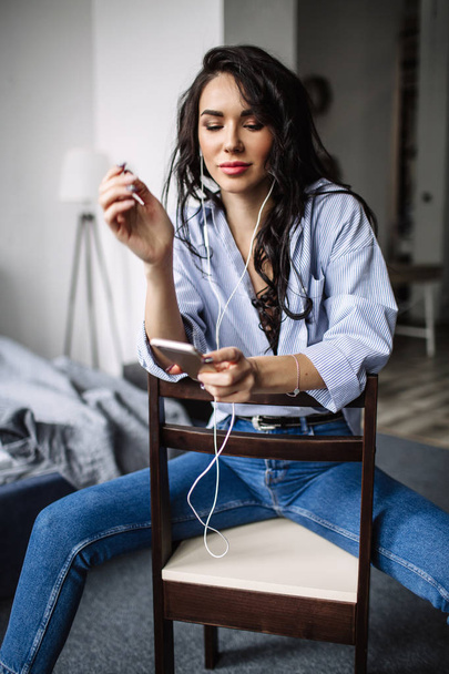 A beautiful brunette listens to music in headphones in a room near the sofa, is dressed in a shirt and blue jeans, blue - 写真・画像