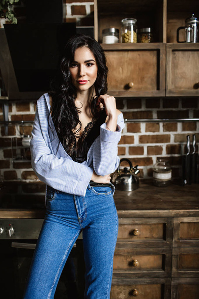 Beautiful brunette in the kitchen, modern housewife, fashion, wooden brown furniture, dressed in blue jeans and a blue shirt - Fotoğraf, Görsel