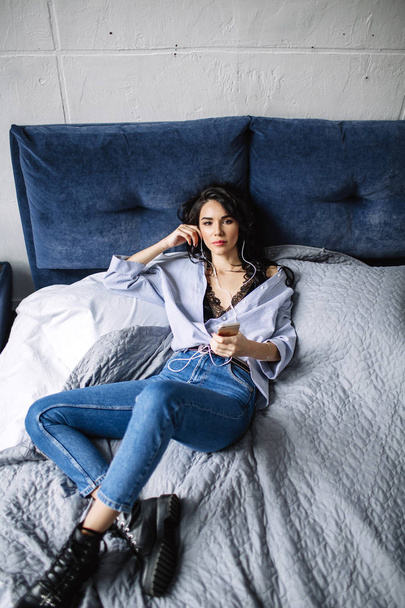 A beautiful brunette listens to music in headphones in a room near the sofa, is dressed in a shirt and blue jeans, blue - Foto, Imagen