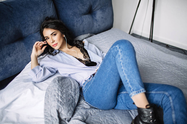 A beautiful brunette listens to music in headphones in a room near the sofa, is dressed in a shirt and blue jeans, blue - Photo, image