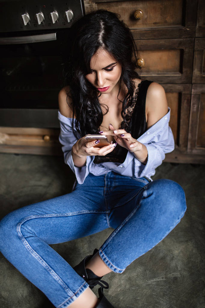 Beautiful brunette in the kitchen, modern housewife, fashion, wooden brown furniture, dressed in blue jeans and a blue shirt - Zdjęcie, obraz