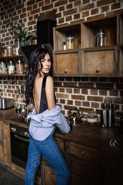 Beautiful brunette in the kitchen, modern housewife, fashion, wooden brown furniture, dressed in blue jeans and a blue shirt - Photo, Image