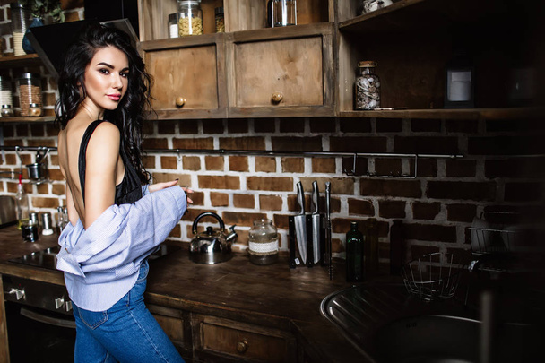 Beautiful brunette in the kitchen, modern housewife, fashion, wooden brown furniture, dressed in blue jeans and a blue shirt - Zdjęcie, obraz