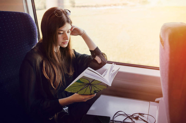 красивая девушка-хипстер, путешествующая на поезде и держащая в руках книгу. стильная счастливая женщина, читающая книгу у иллюминатора в поезде. путешествия и транспорт. пространство для текста
 - Фото, изображение