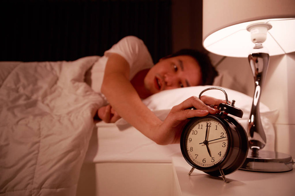 朝は目覚まし時計に手を拡張するとベッドで眠そうな若い男 - 写真・画像