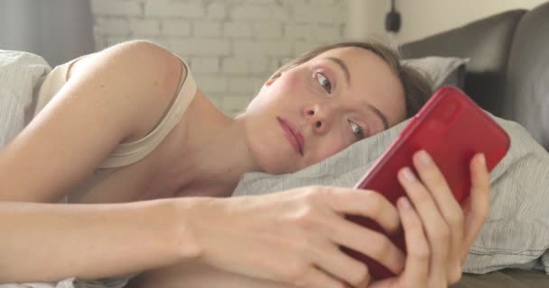 自宅の寝室のベッドでスマート フォンで女性のテキスト メッセージ - 映像、動画