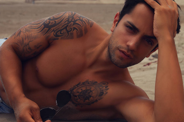 Foto van een knappe man liggend op het strand houden zonnebril, kijken naar de camera. - Foto, afbeelding