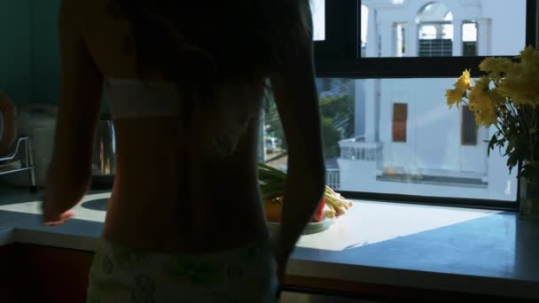 backside view young woman in shorts shifts green onion from bell peppers to tomatoes against window - 映像、動画