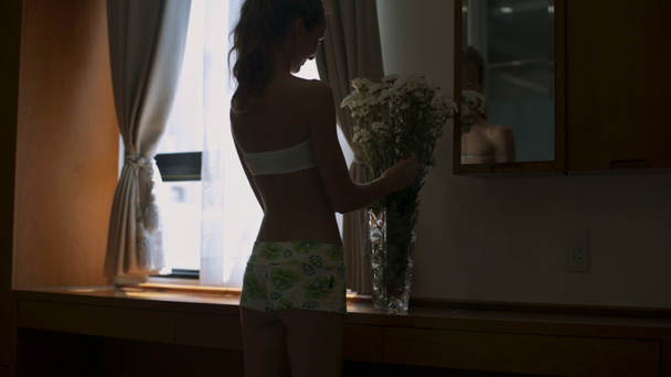 backside view pretty girl in shorts comes to flower in vase on windowsill looks at mirror reflection and smooths hair - Felvétel, videó