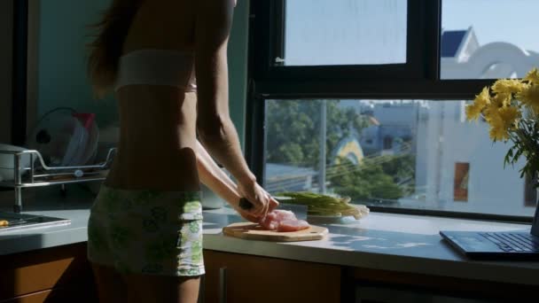 long haired girl silhouette cuts meat into pieces on white kitchen table with yellow flowers in front of window  - Felvétel, videó