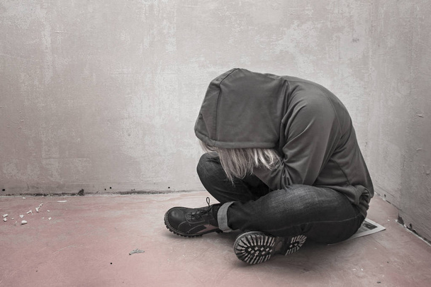 Foto eines verzweifelten jungen Drogenabhängigen, der Kapuze trägt und allein in der Ecke sitzt - Foto, Bild